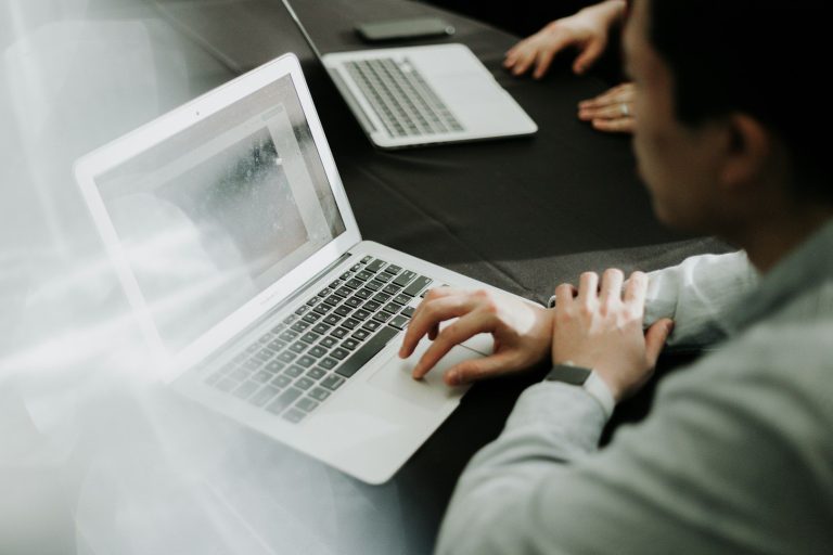 Persoon aan tafel met laptop, bezig met werkzaamheden. Een tweede laptop zichtbaar.
